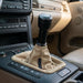 Close-up of a manual gear shift knob with a beige fabric boot inside a car's center console, showing beige leather seats and dashboard.