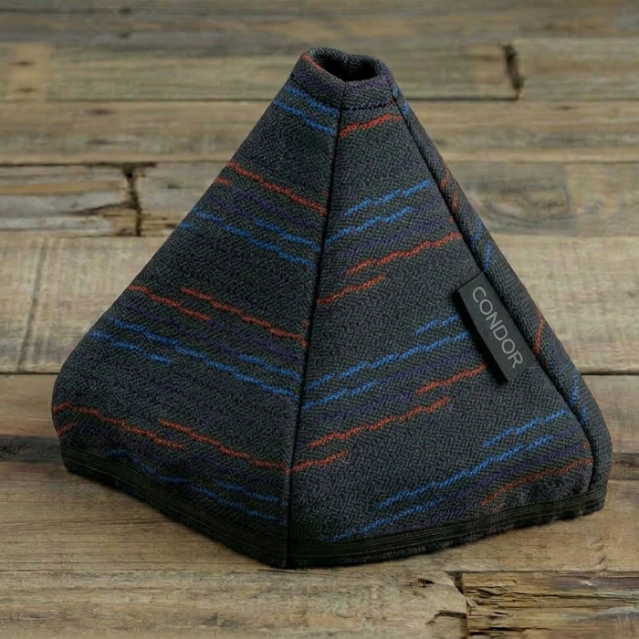 A triangular fabric cover with a dark gray base and thin blue and orange stripes, resting on a rustic wooden plank surface. A small black fabric tag on the side reads 'COOLGIRL'.