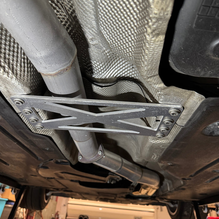 A close-up underside view of a vehicle showing a silver metal undercarriage support bracket mounted near the exhaust pipe, with the garage floor and another vehicle visible in the background.