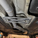 A close-up underside view of a vehicle showing a silver metal undercarriage support bracket mounted near the exhaust pipe, with the garage floor and another vehicle visible in the background.