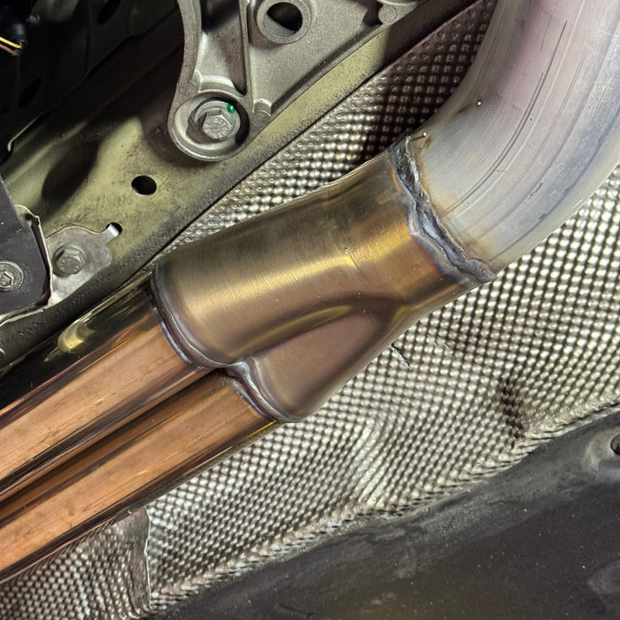 Close-up of a welded metal exhaust pipe fitting with copper-colored tubes, shown installed against a textured vehicle underbody panel.
