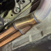 Close-up of a welded metal exhaust pipe fitting with copper-colored tubes, shown installed against a textured vehicle underbody panel.