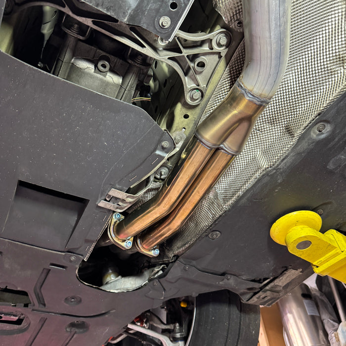 A close-up view of a vehicle's undercarriage showing a custom exhaust system with dual copper-colored pipes, a silver metal pipe, and a heat shield, mounted on a vehicle frame.